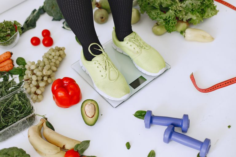 Elevated Mind, Body & Life article image of a person standing on scales surrounded by fresh, low calorie dense produce and fitness equipment, symbolizing a balanced lifestyle.