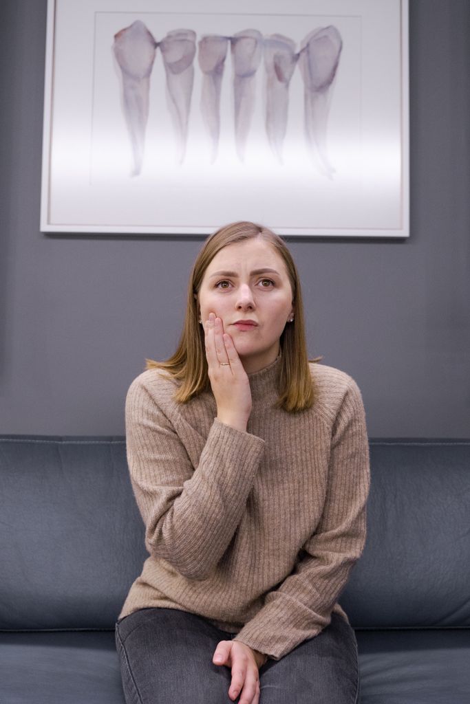 Woman in a brown sweater holding her cheek, indicating oral problems, sitting in a dental clinic setting.